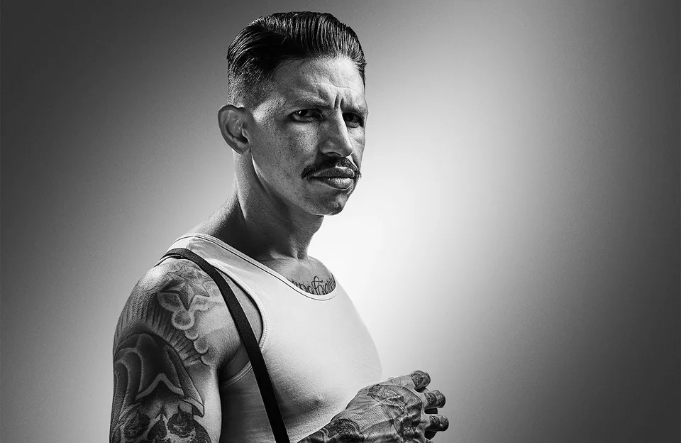 Barbeiro Willy Morales em retrato preto e branco, de bigode, regata, suspensórios e braços tatuados